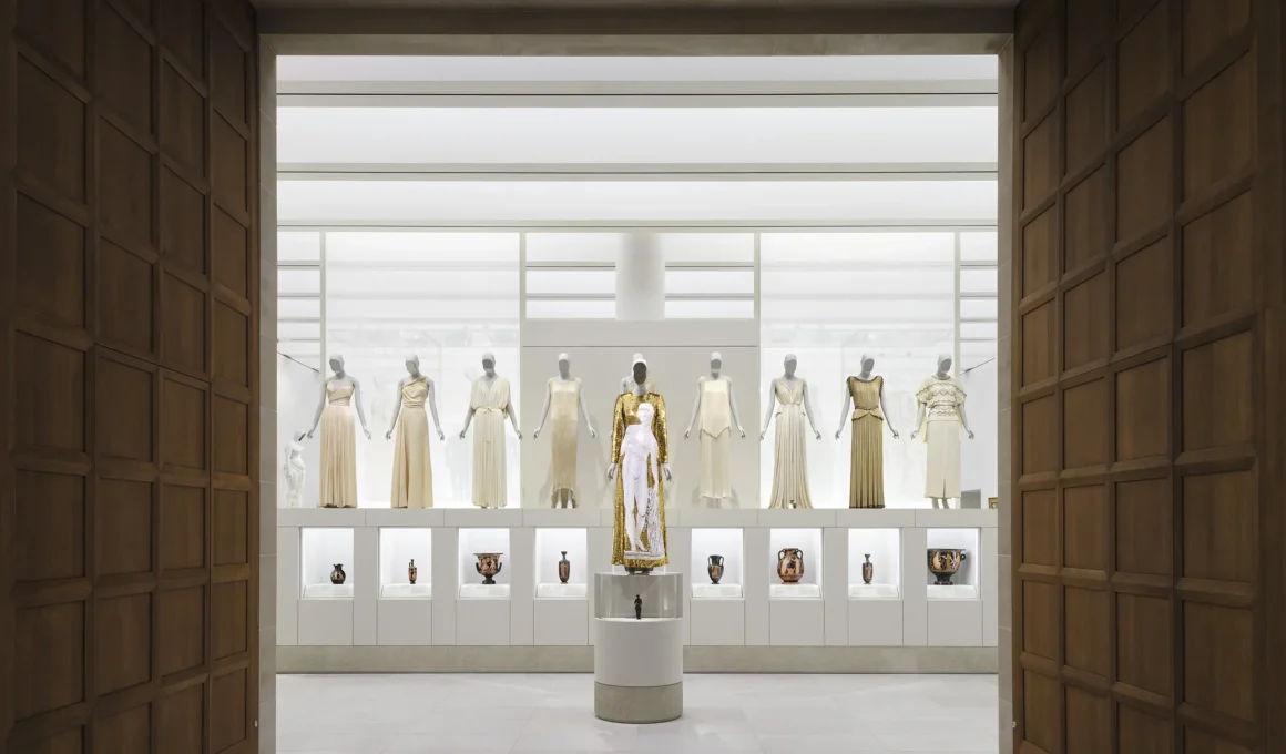 Long spatial view of a high-ceiling museum gallery showing classical sculpture juxtaposed with contemporary mannequin displays on low platforms under soft indirect lighting.