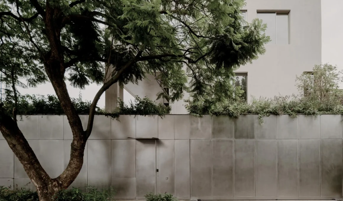 Brutalist concrete exterior facade of a Mexico City house behind a metallic gate and mature green trees.