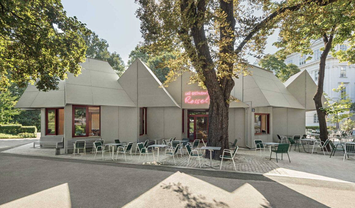 This eye-level shot captures the building's cluster-like form, appearing as several small houses facing the park. Outdoor tables and green chairs are arranged on a paved area beneath mature trees, highlighting the project's commitment to tree preservation.