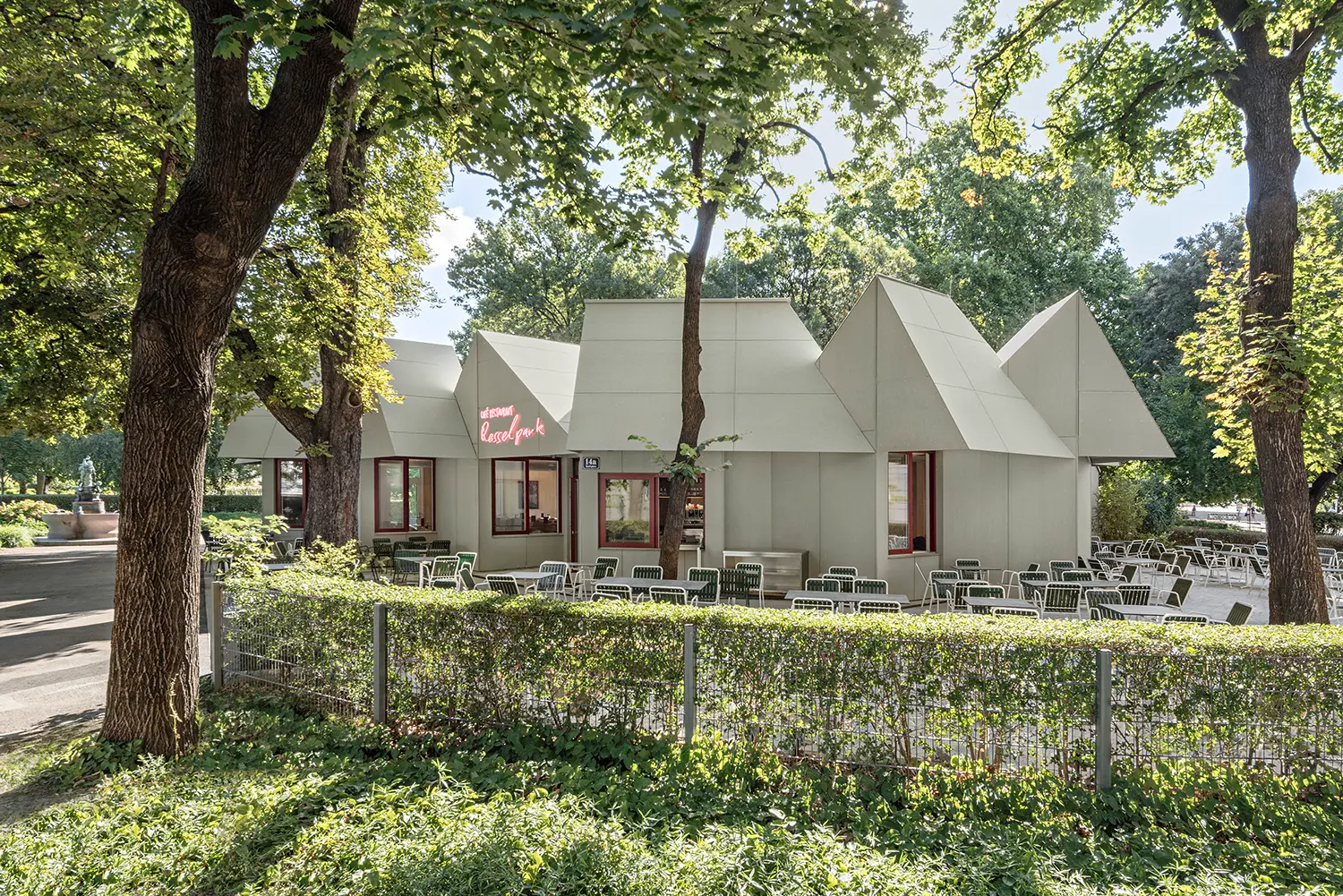 The image shows an interior corner niche with built-in wooden benches and light-colored tables. A large sliding window offers views of the park, while a slanted ceiling with acoustic panels emphasizes the individual "house fragment" geometry.