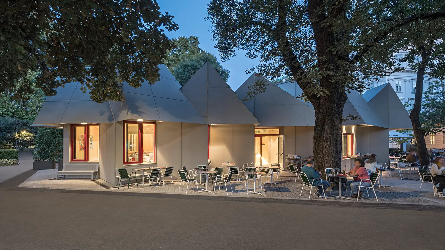 Dusk view of Café Restaurant Resselpark with illuminated windows and a neon sign in the park.