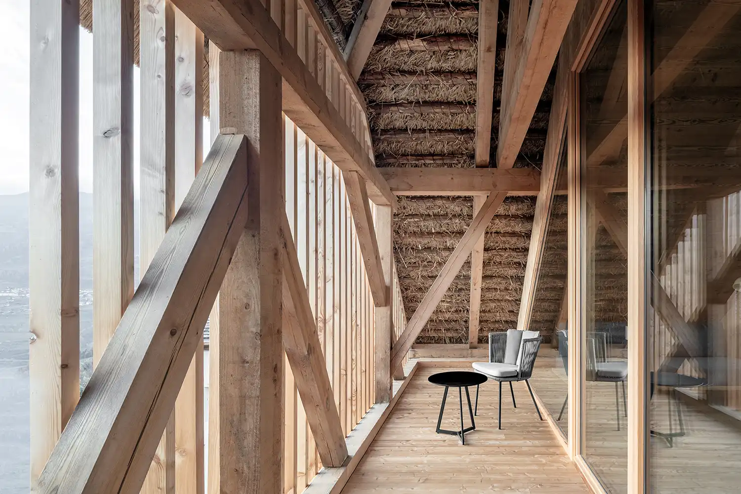Interior view of a wooden balcony with exposed structural timber beams and a view of the Laion valley through glass.