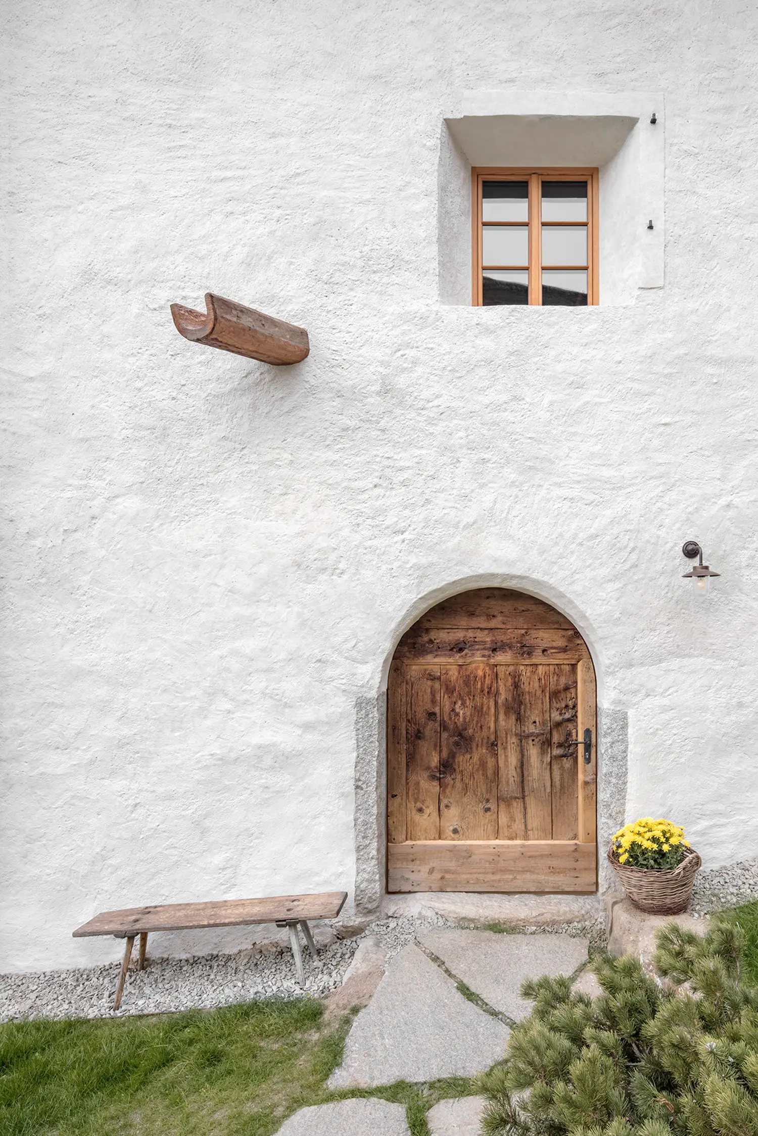 White textured plaster wall with a small wooden window, a timber waterspout, and an arched wooden door at the base.
