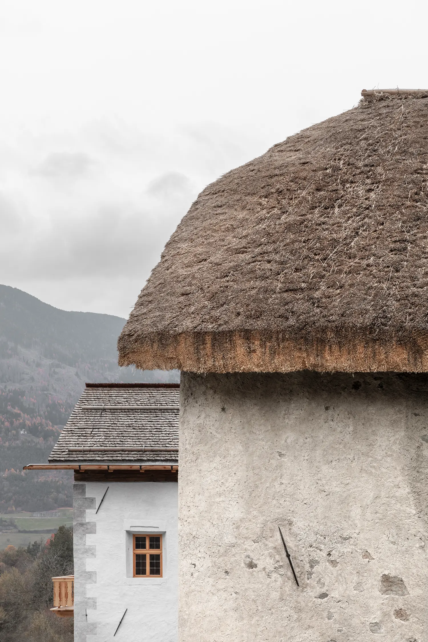 External view of two Alpine buildings with contrasting thatched and shingled roofs against a cloudy mountain backdrop.