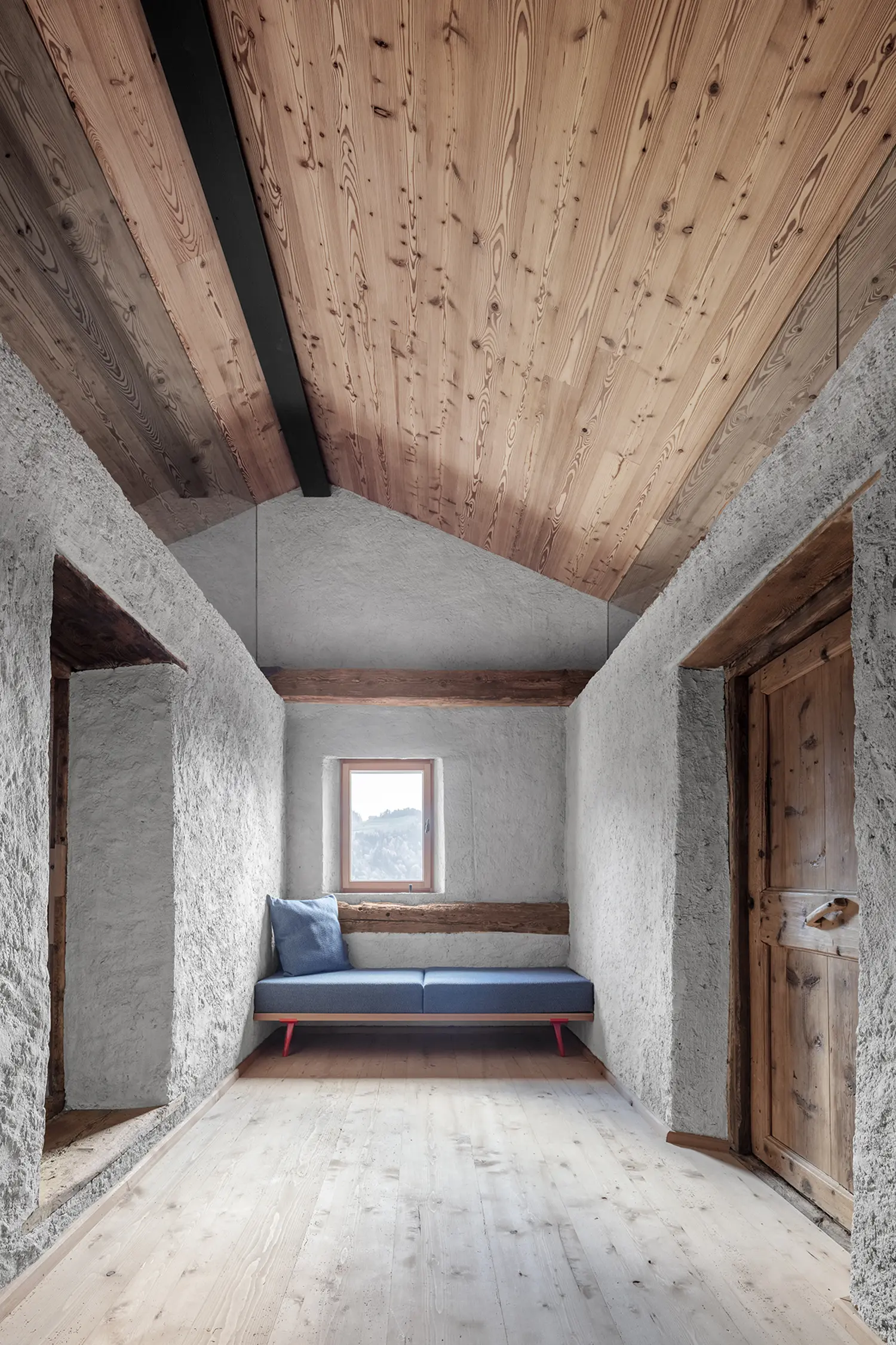 Narrow interior hallway with rough-textured white walls, a timber ceiling, and a blue daybed under a small window.
