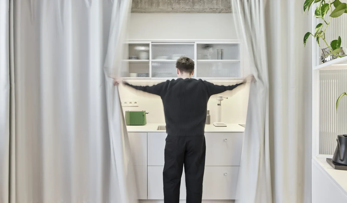 A person standing with arms outstretched opening floor-to-ceiling white curtains to reveal a minimalist white kitchen counter and shelving units.