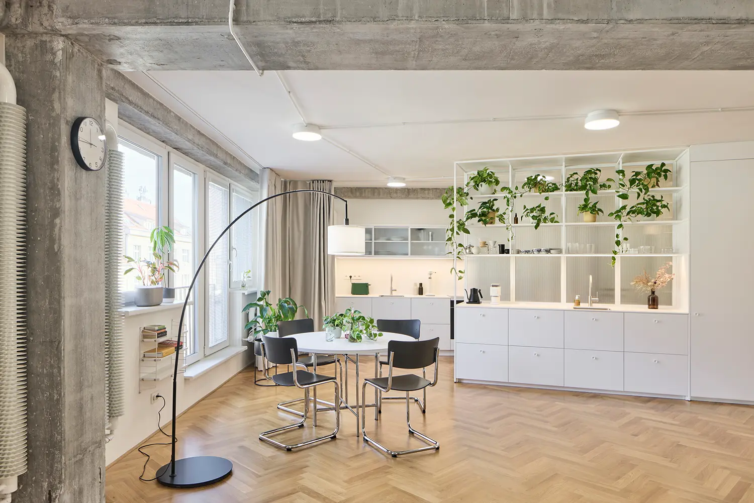 Open-plan living and dining room with herringbone wood flooring, a circular white dining table, and a large white shelving divider unit filled with green plants.