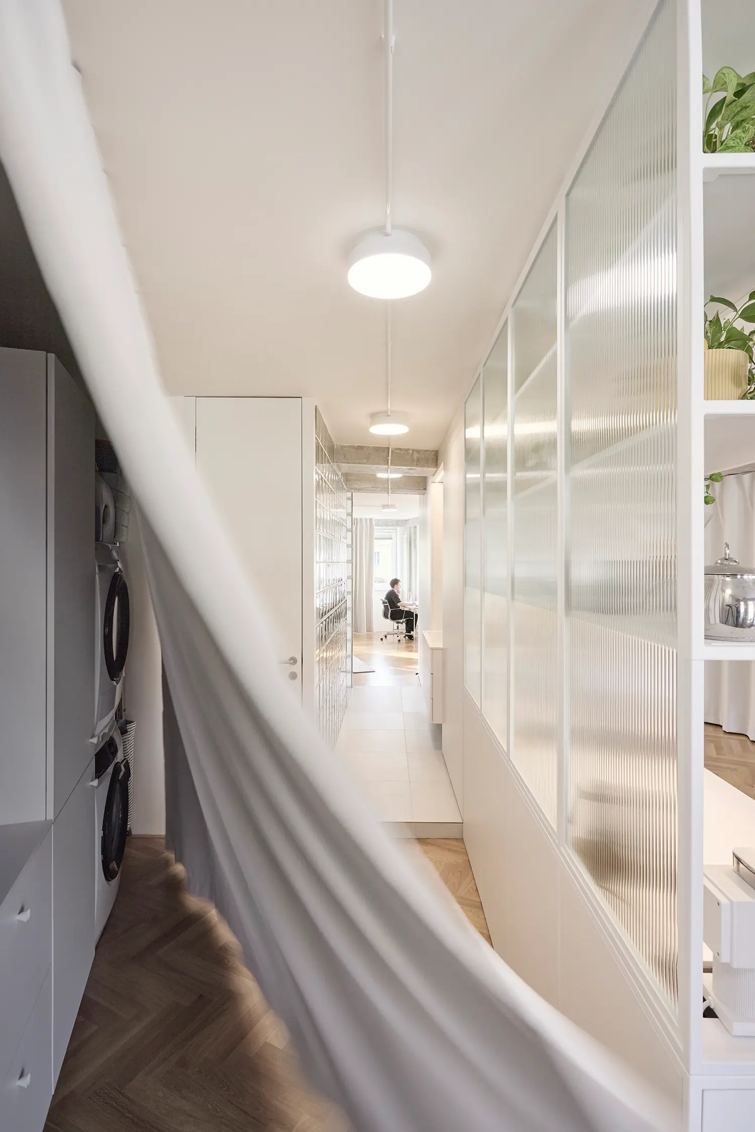 A long white hallway flanked by a translucent fluted glass wall on one side and a sweeping white curtain captured in motion on the other.