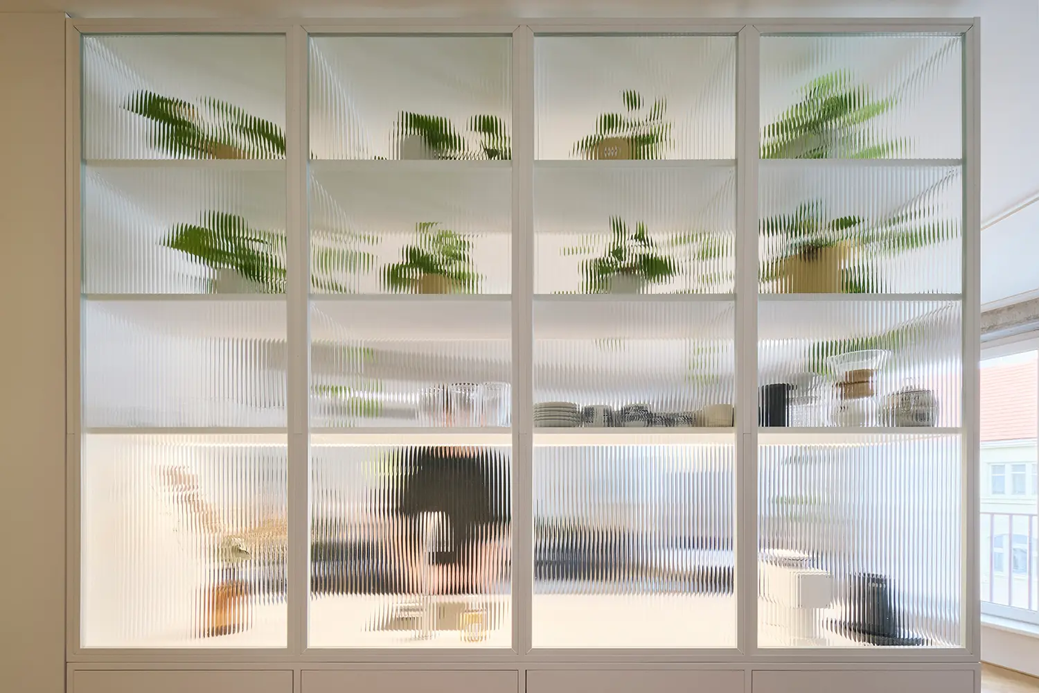 Close-up of a white partition wall made of fluted translucent glass panels displaying silhouettes of green plants on the shelves inside.