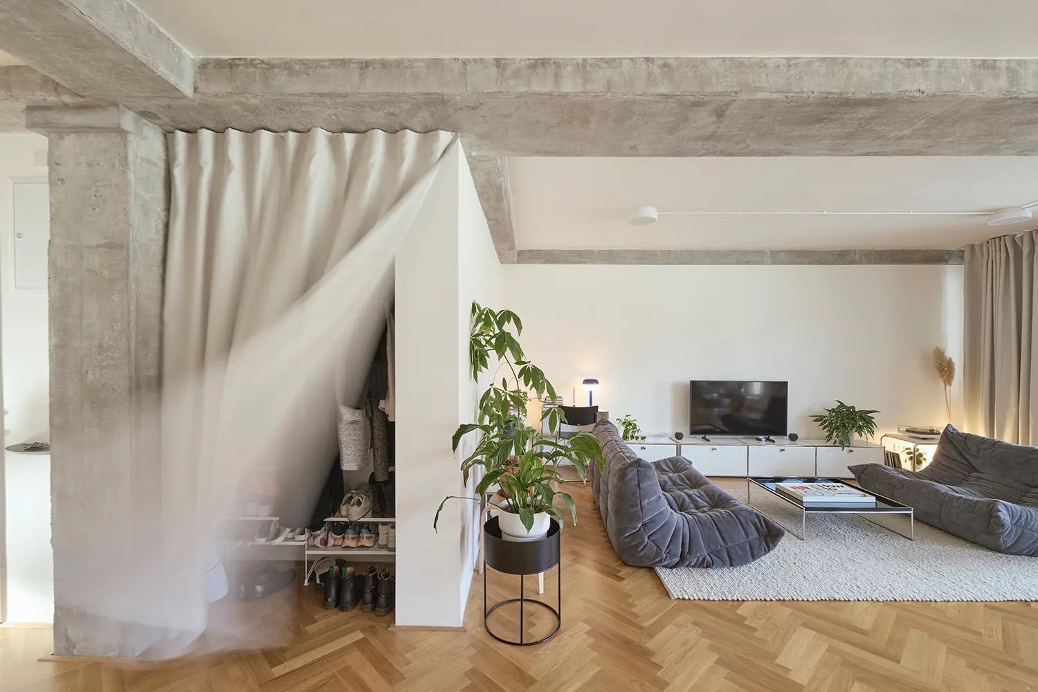 Living room with grey modular sofas on a textured rug under an exposed concrete beam, featuring a white curtain partially drawn back to reveal storage.