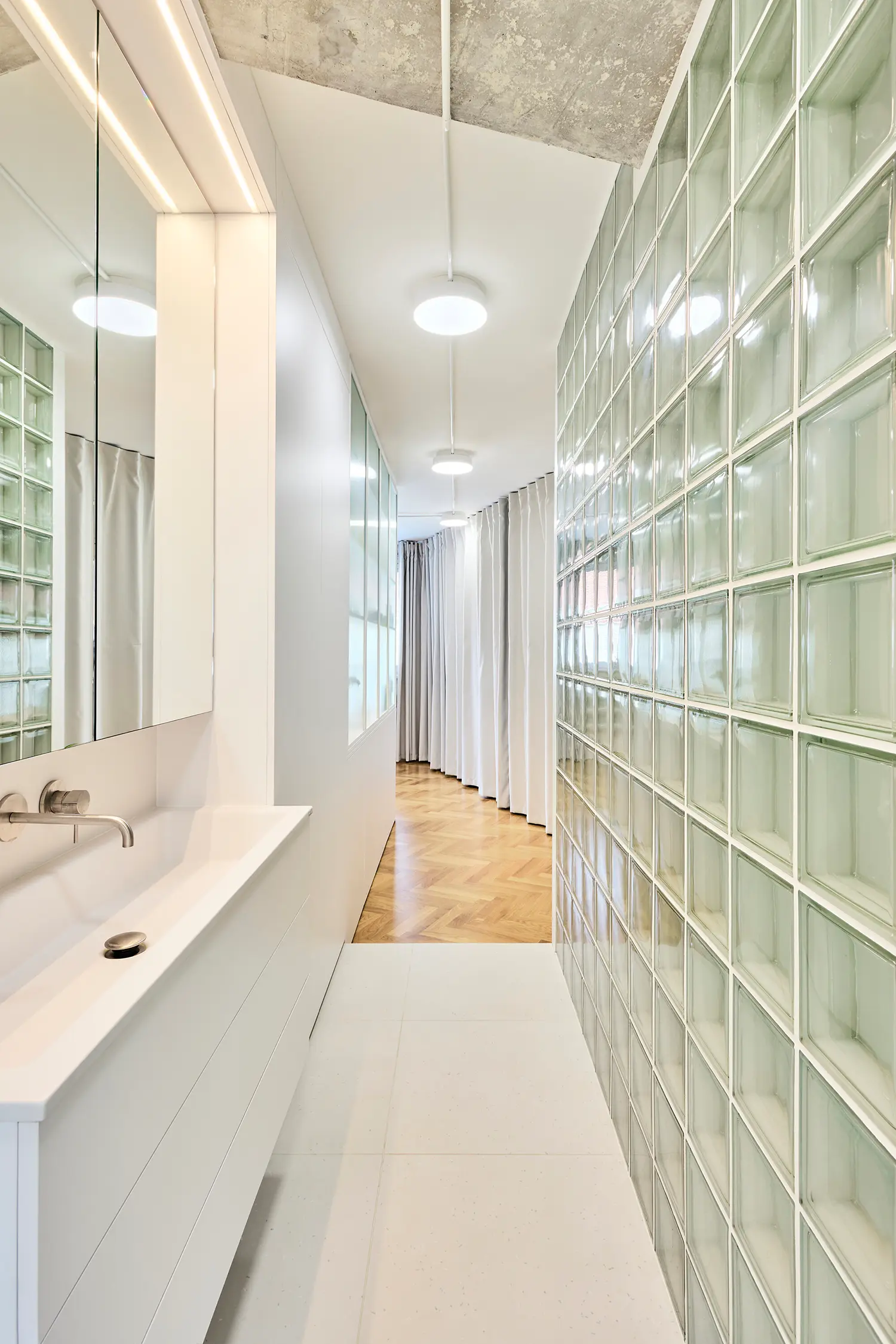 A bright white bathroom corridor with a long modern sink vanity on the left and a translucent glass block wall on the right.