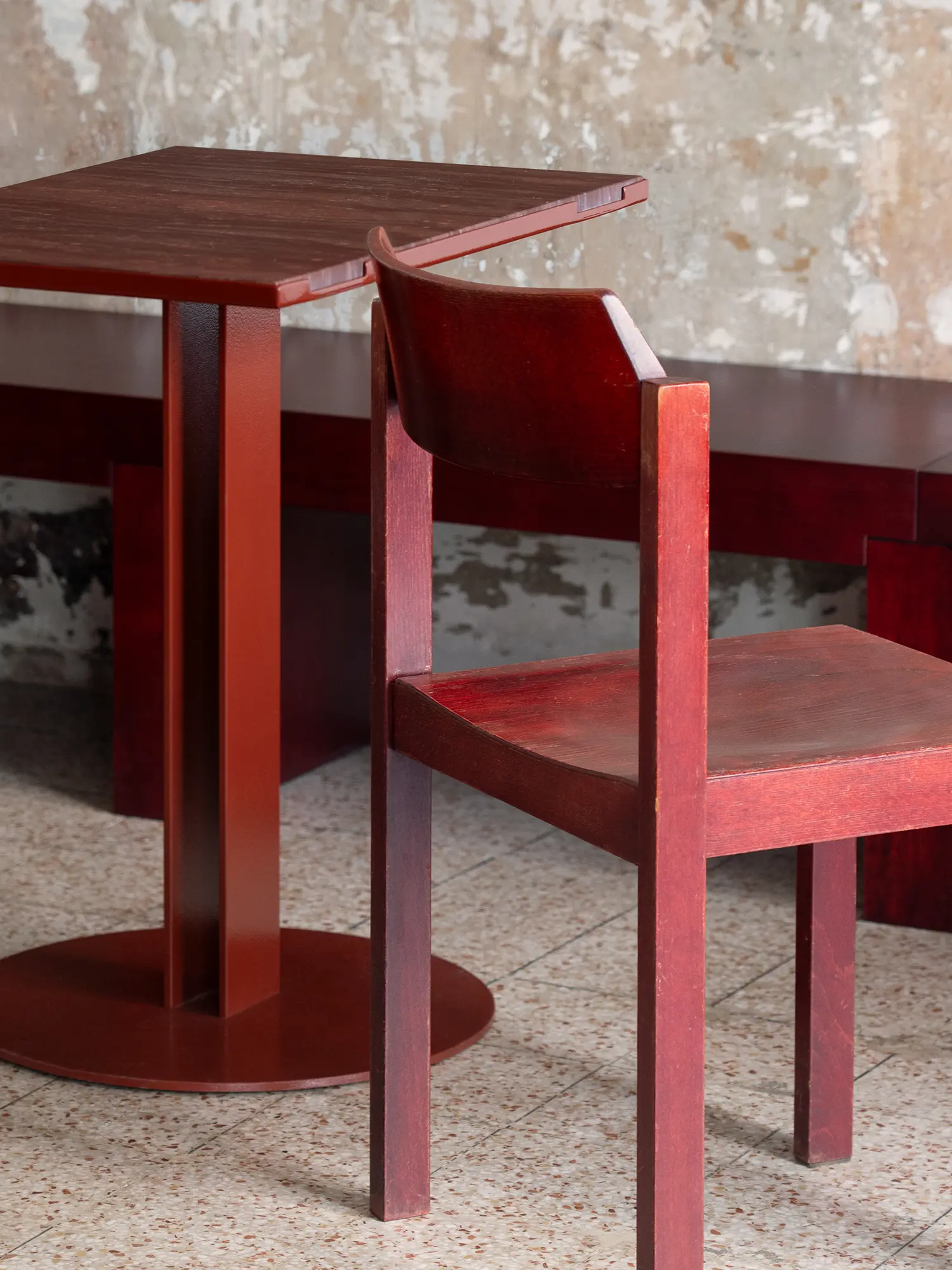 Detail of a red custom table with orange-peel texture and a vintage wooden chair against a weathered wall.