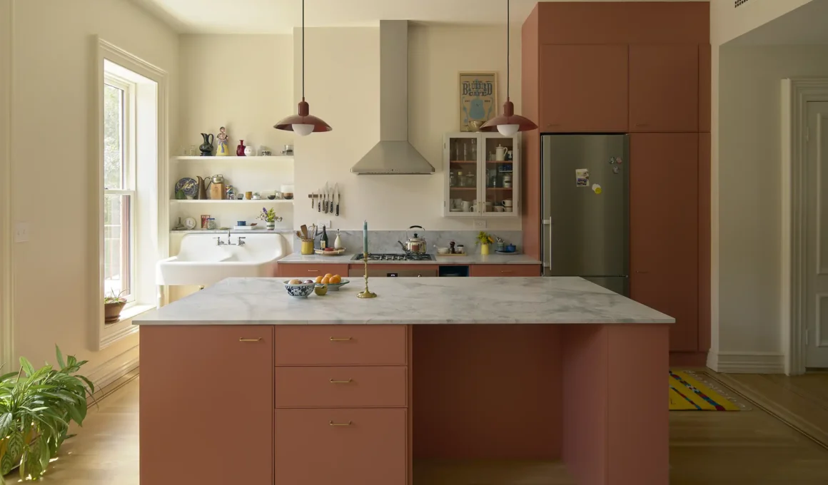 Modern kitchen with terracotta-colored cabinetry, a marble island, and a vintage white farmhouse sink under a window.