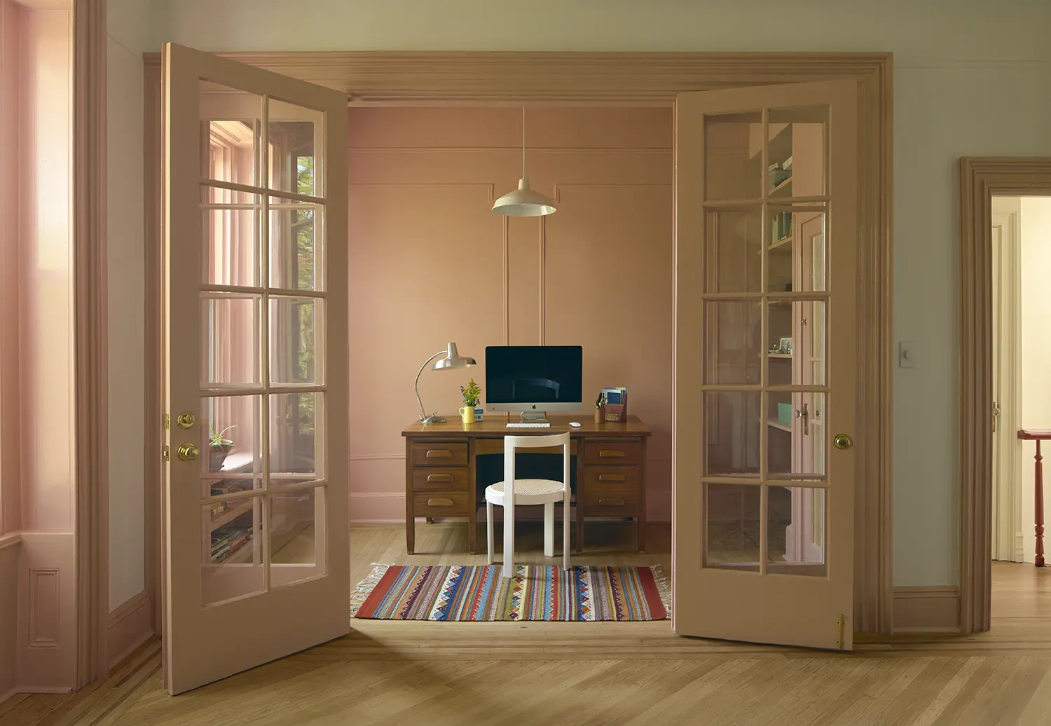 View through open double French doors into a peach-colored home office with a classic wooden desk and a modern computer.