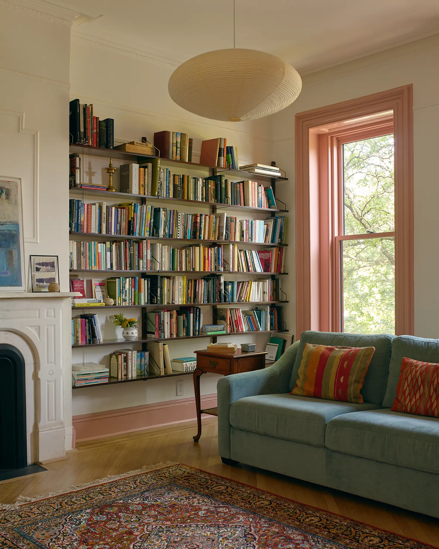 Living room with floor-to-ceiling built-in bookshelves, a white marble fireplace, and a light blue sofa near a pink-trimmed window.