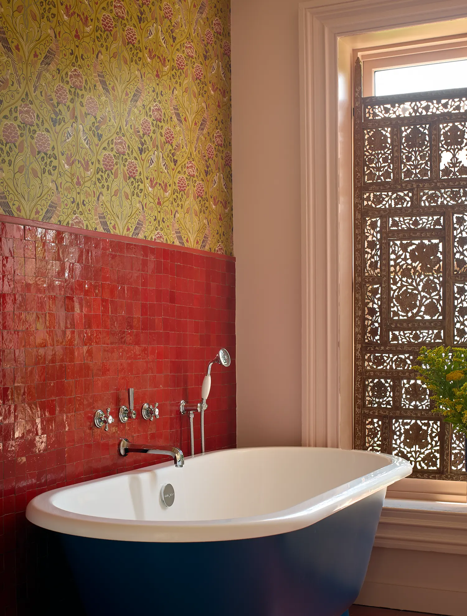 A blue-based clawfoot bathtub set against a wall of bright red square tiles and intricate floral wallpaper.