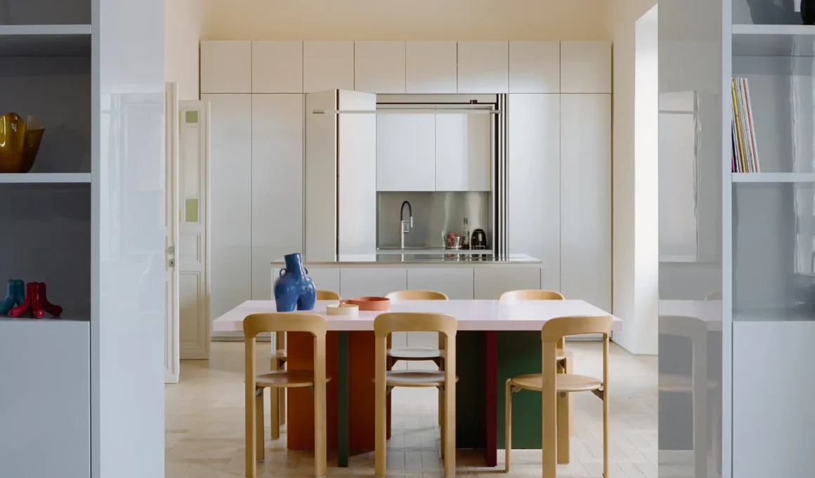 Open-plan dining room with colorful table, light wood parquet floor, and recessed kitchen cabinet by Set Architects.