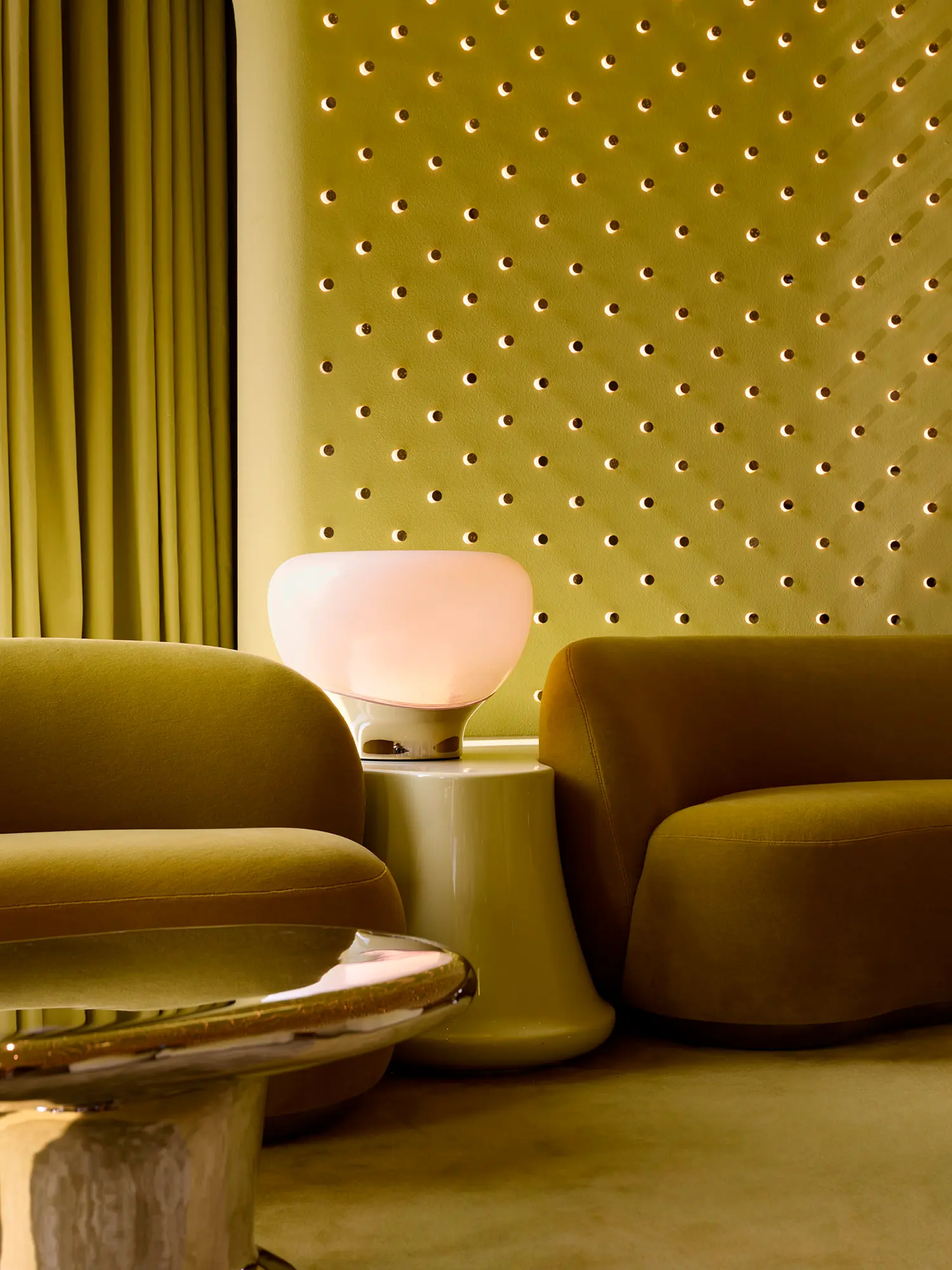 Soft mohair armchairs and a glowing mushroom-shaped table lamp against a green textured wall.
