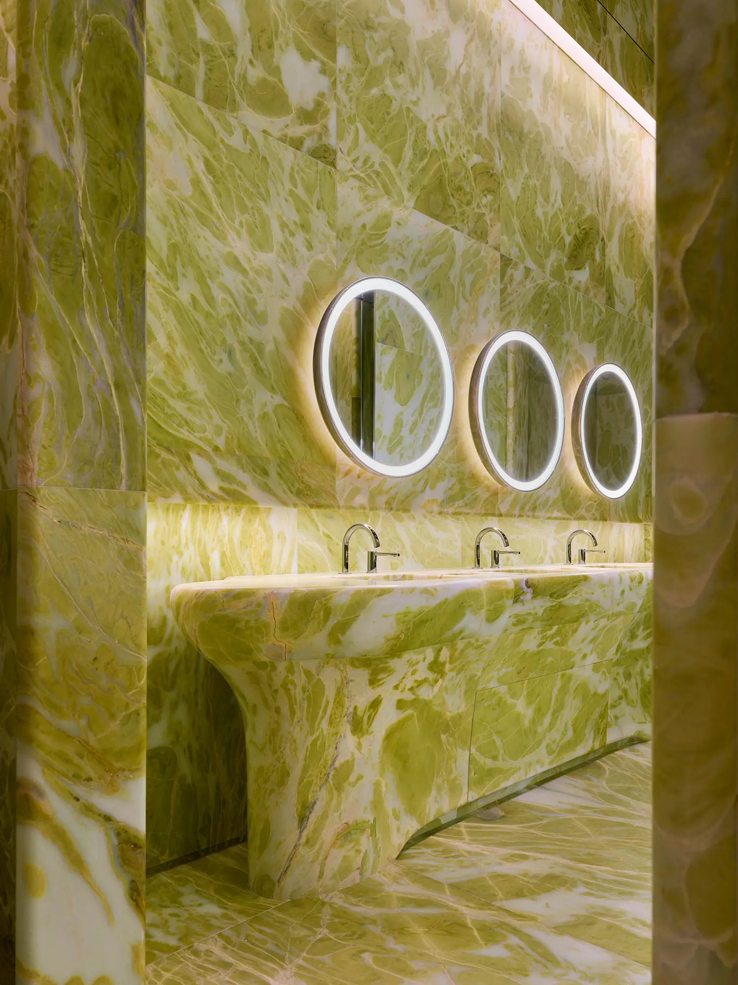 Luxurious green marble restroom with integrated carved sinks and circular backlit mirrors.