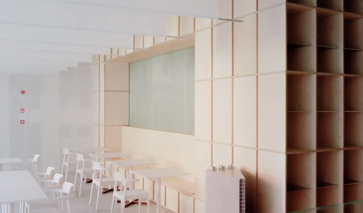 Light birch plywood modular shelving and wall panels next to minimalist white dining tables and chairs in a contemporary cafe interior.