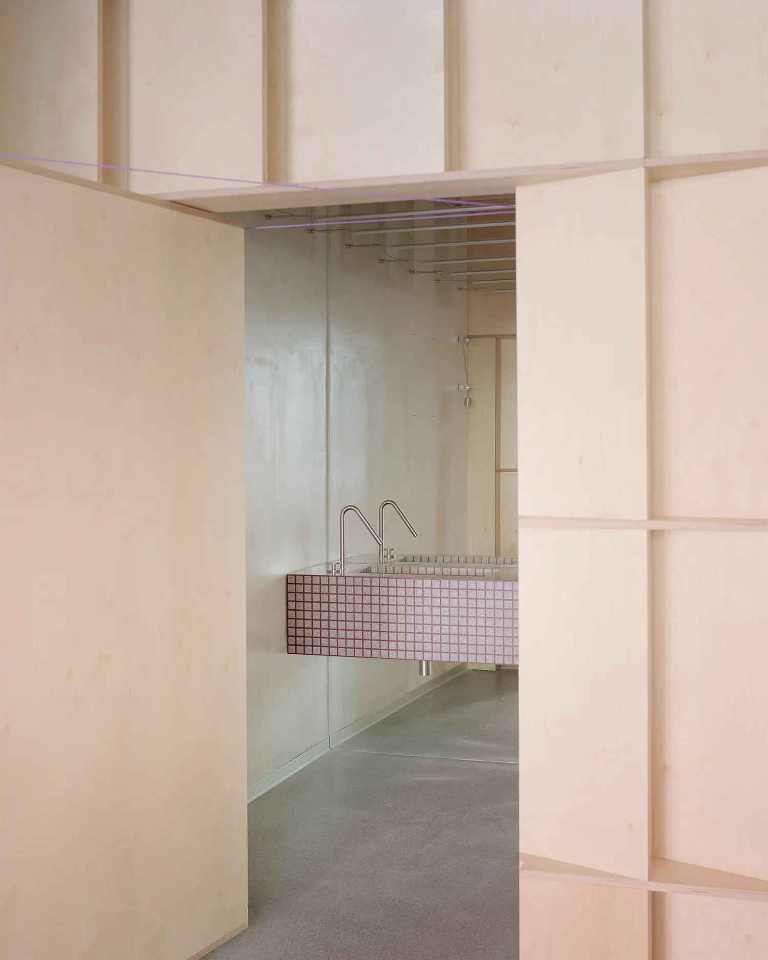 View through an open birch wood doorway showing a modern bathroom sink clad in pink gridded tiles with minimalist chrome faucets.