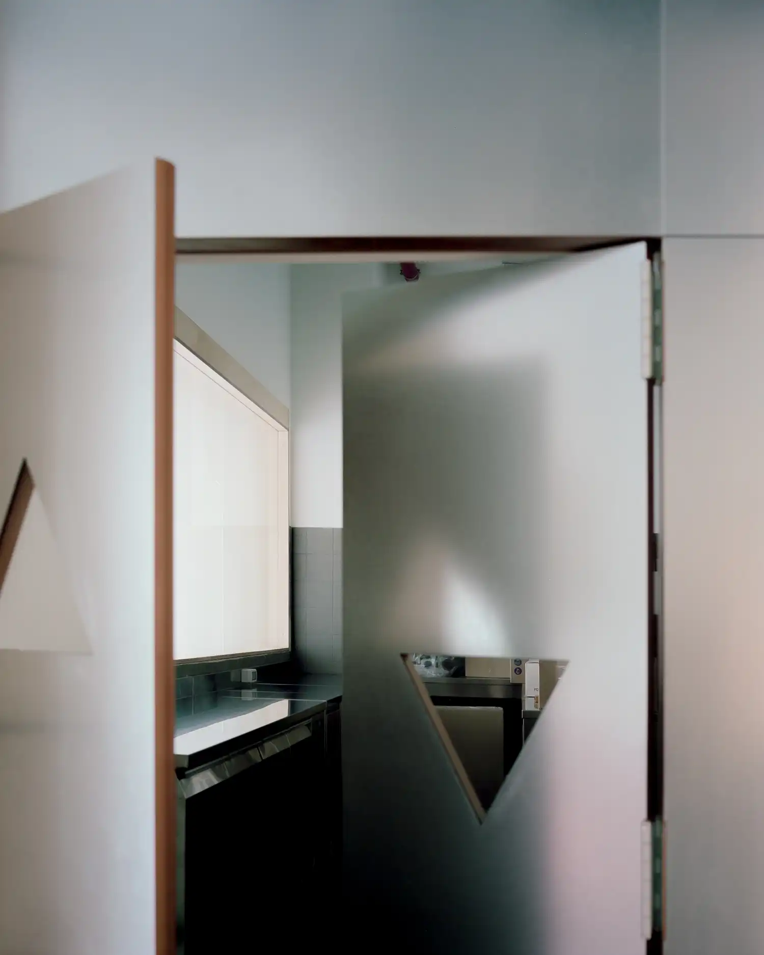 View through custom metal double doors with triangular cutouts into a professional cafe kitchen featuring stainless steel counters and a large glowing frosted light panel.