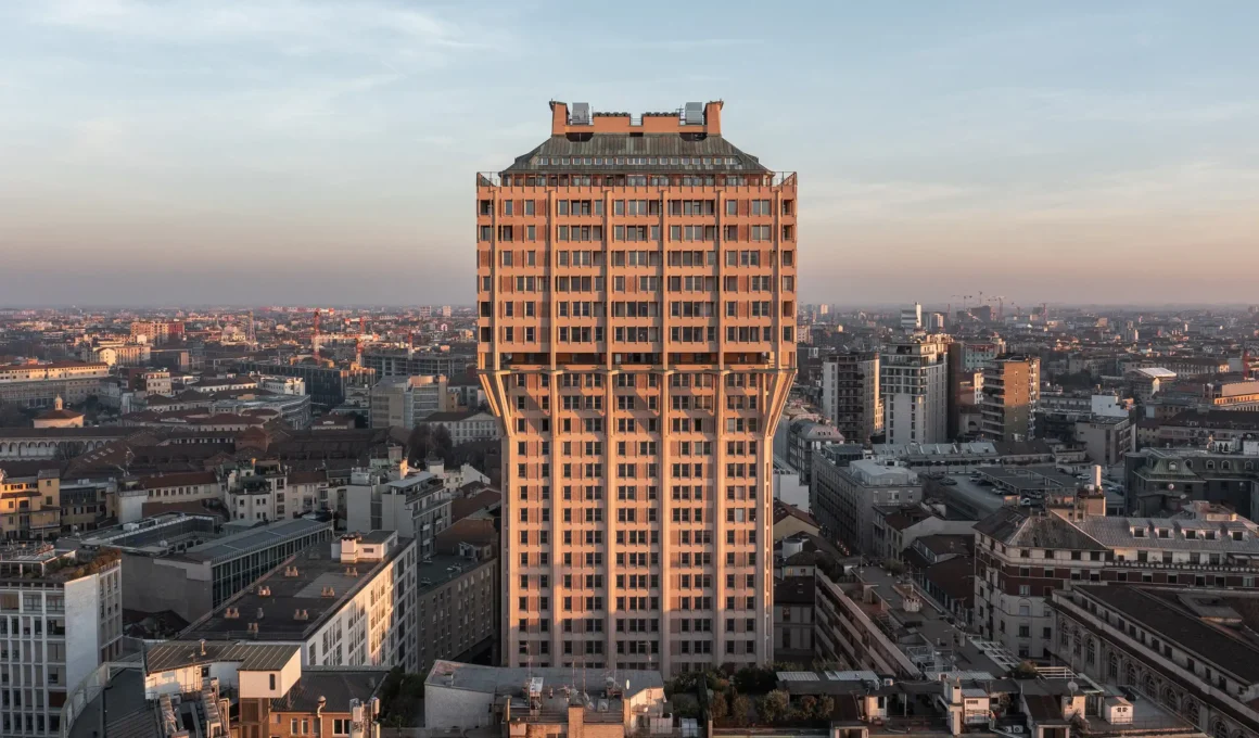 A symmetrical, front-facing aerial photograph of the Brutalist Torre Velasca in Milan, bathed in the warm, golden light of a setting sun against a pastel sky.
