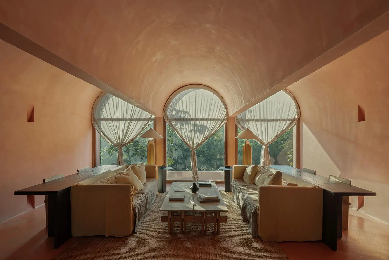 Interior of a living room with a high vaulted ceiling, Chukum walls, and a large arched window overlooking trees.