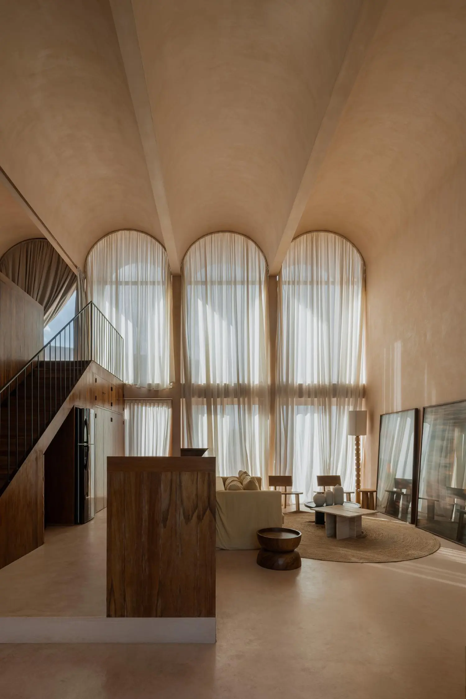 Double-height interior loft with a mezzanine, wooden carpentry, and triple arched windows with sheer curtains.