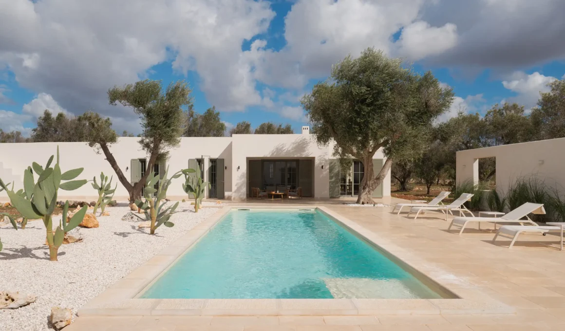 Rectangular swimming pool reflecting a cloudy blue sky, flanked by white sun loungers and prickly pear cacti against a minimalist white villa.