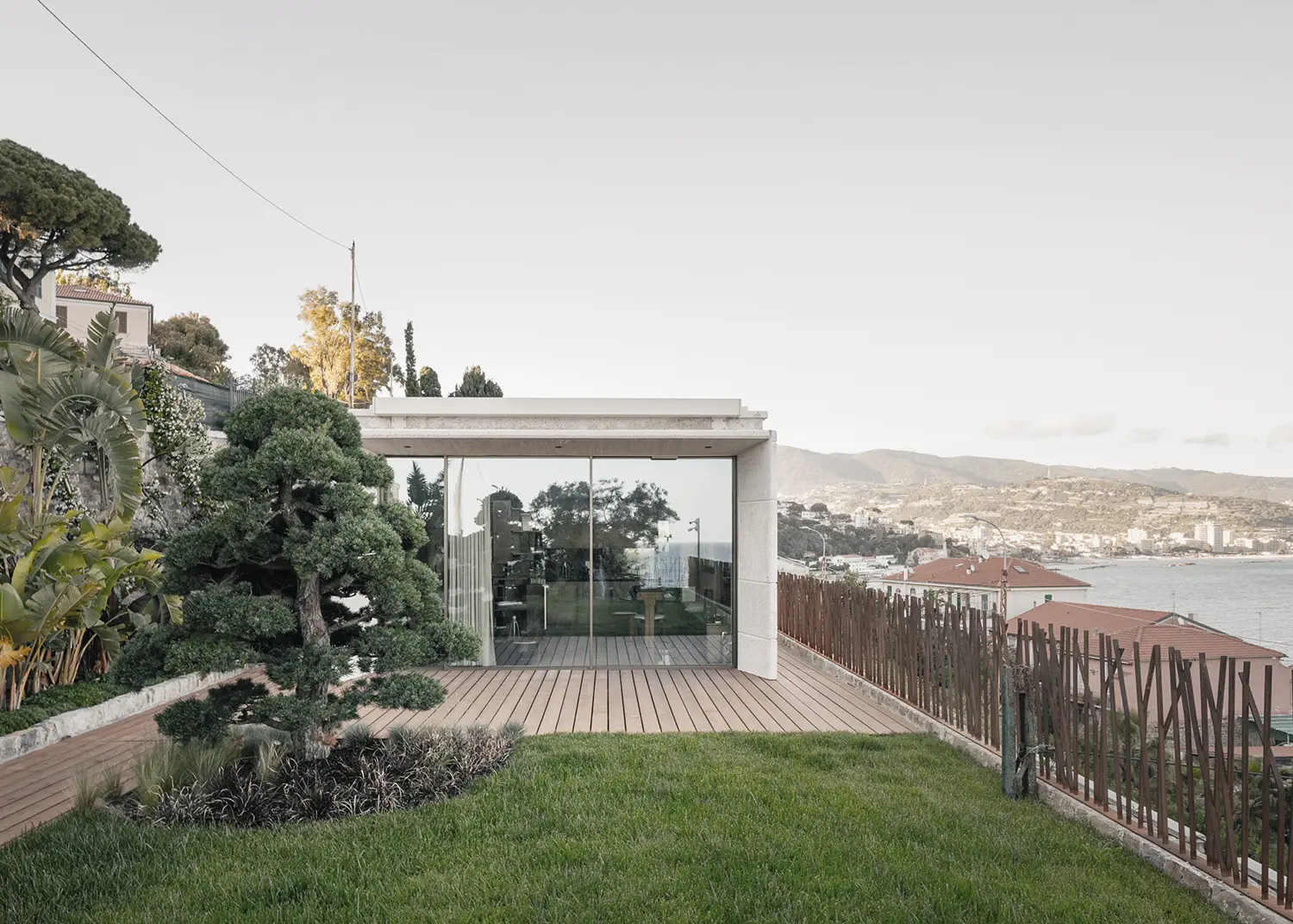 Villa S-2113 Calvi Ceschia Viganò, exposed concrete volume, full-height glazing, timber deck, coastal garden with sculpted pine