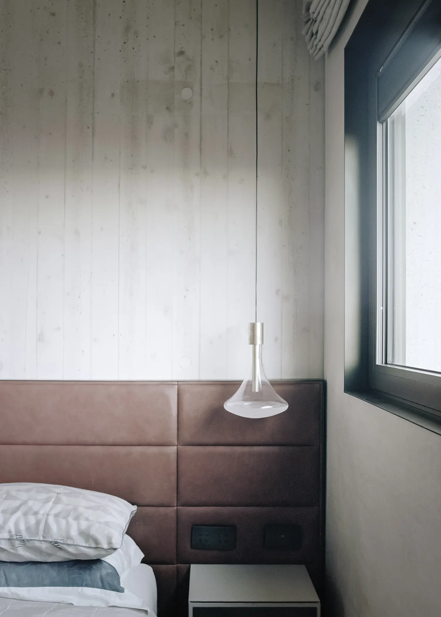 Villa S-2113 Calvi Ceschia Viganò, bedroom with board-formed concrete wall, leather upholstered headboard, glass pendant lamp