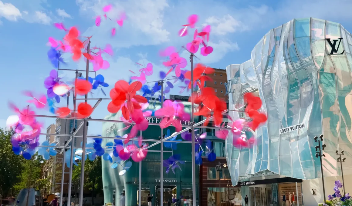 Vincent Leroy Point Cloud kinetic installation featuring blue, pink, and red translucent discs on a metal frame at Taikoo Li Sanlitun in Beijing, with glass luxury boutique storefronts under a blue sky.