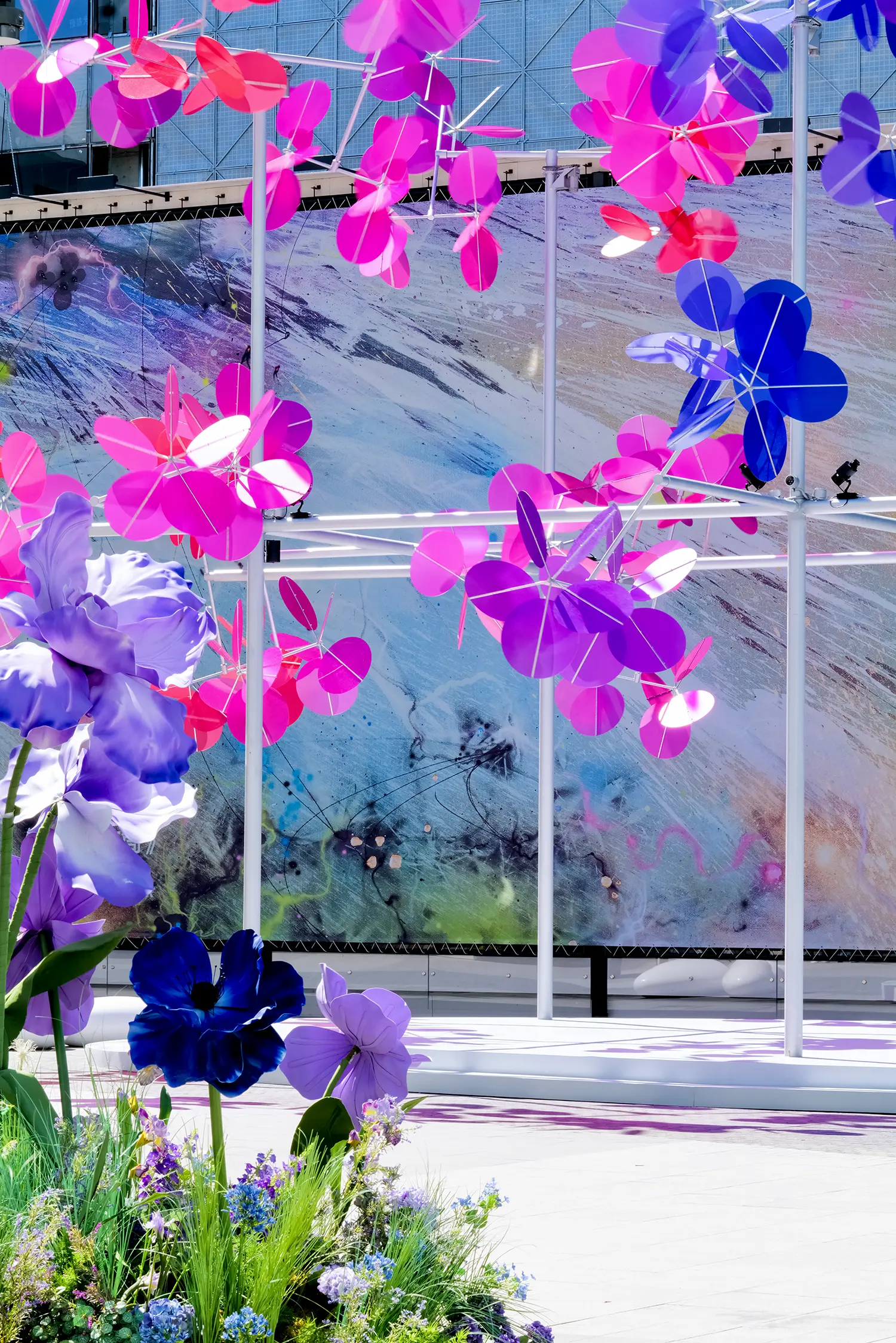 Close-up of Vincent Leroy Point Cloud pink and purple discs on a metal frame, contrasted with oversized fabric flowers in the foreground and a colorful graphic billboard behind.