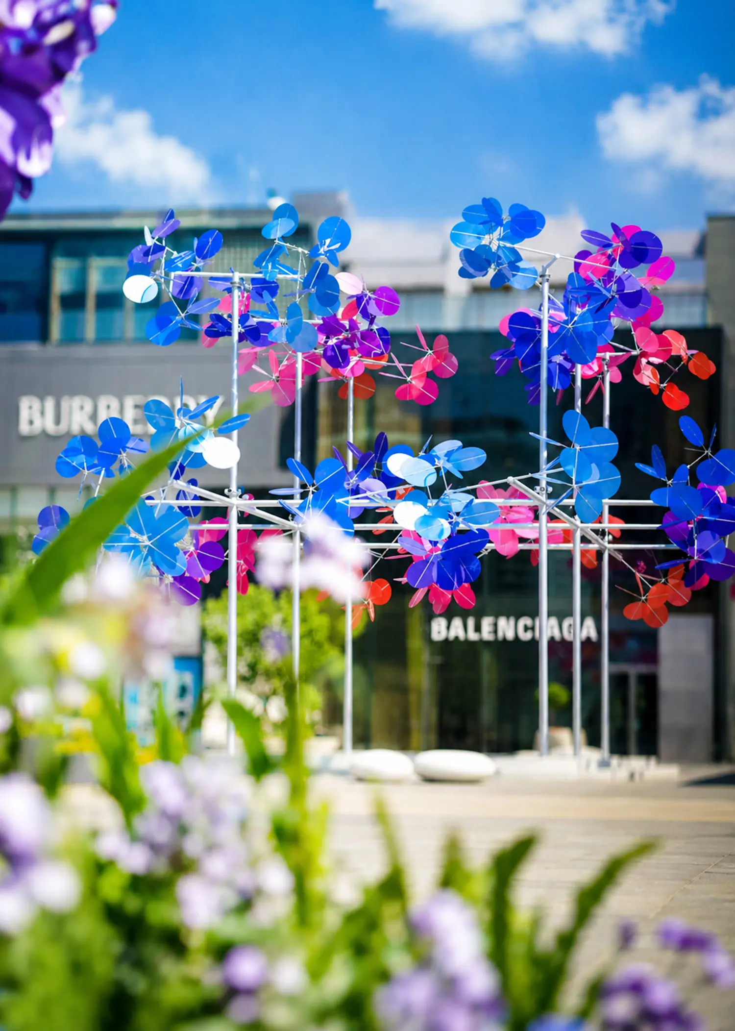 Kinetic sculpture Point Cloud by Vincent Leroy with bright blue and pink discs in front of a Burberry and Balenciaga storefront, blurred purple flowers in the foreground.