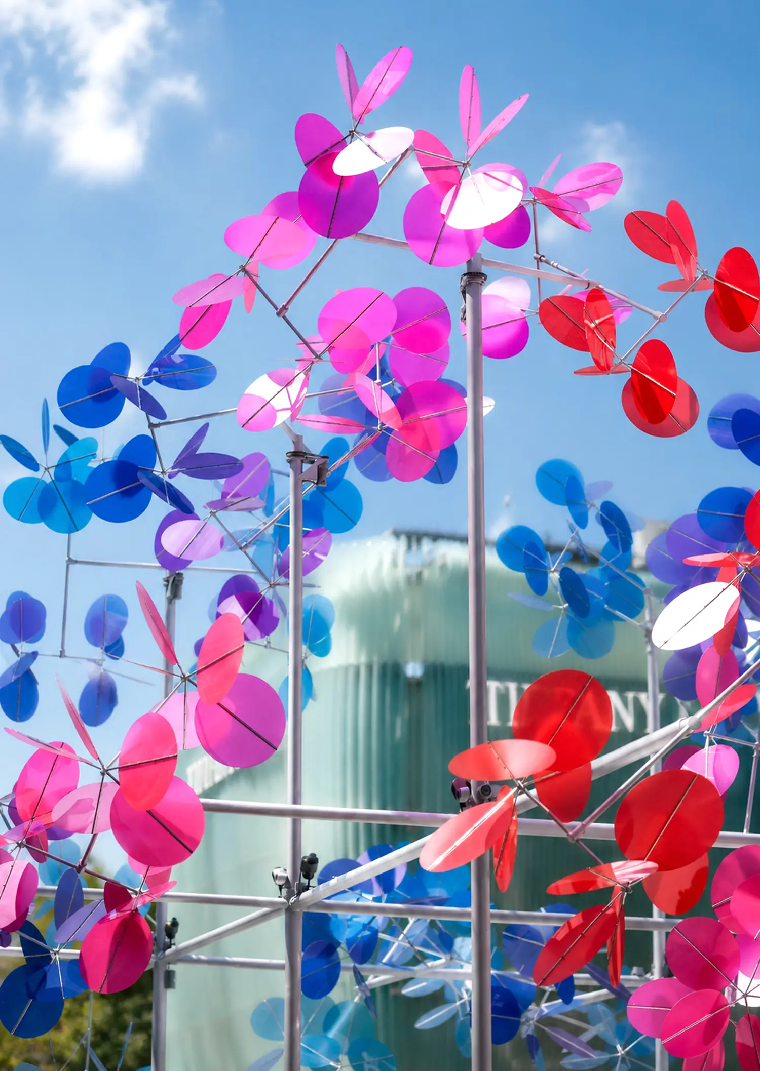 Detail of Vincent Leroy Point Cloud sculpture showing red, pink, and blue translucent discs on a white metal structure with a frosted glass building in the background.