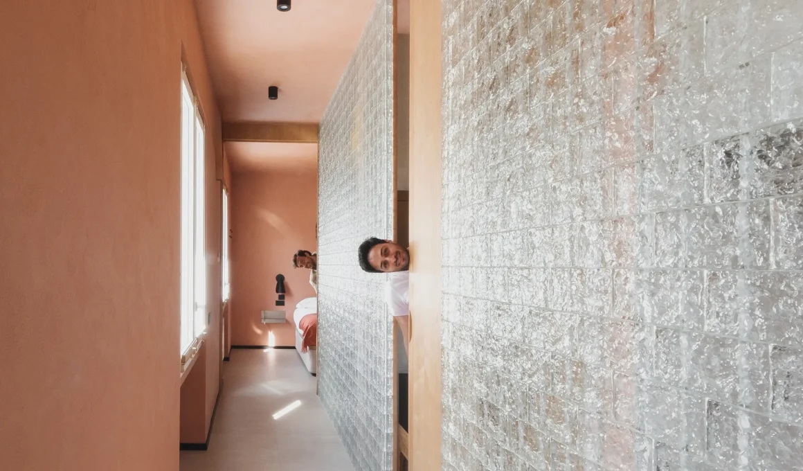 Casa Terra e Vidru by Federico Cartamantiglia, Cagliari — corridor with full-height glass brick wall, terracotta lime plaster walls and ceiling, man peering through gap, ash wood panel