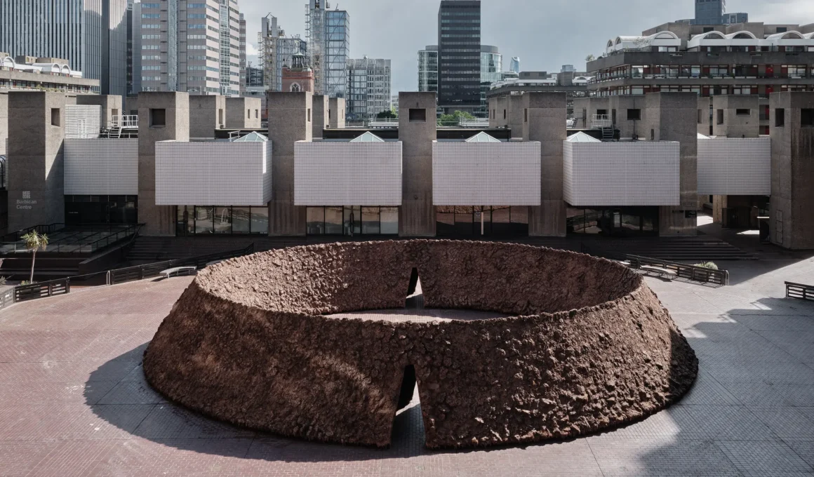 Delcy Morelos origo Barbican Sculpture Court London, aerial view oval earthwork clay soil, brutalist concrete buildings and City of London skyline