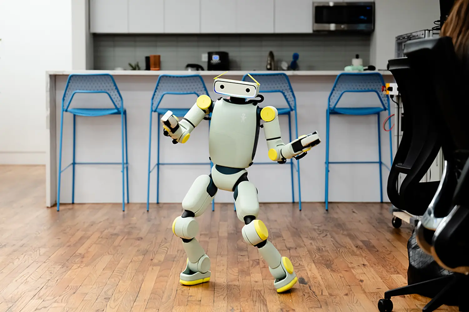 Fauna Robotics Sprout humanoid robot dancing in office kitchen, mint-green and yellow body, bipedal locomotion, blue bar stools background