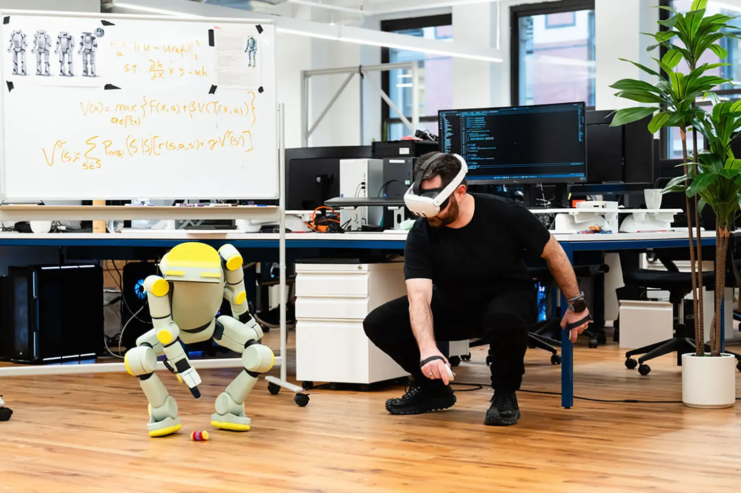 Fauna Robotics Sprout humanoid robot VR teleoperation session, developer in Meta Quest 3 headset crouching alongside robot, office research lab, whiteboard equations