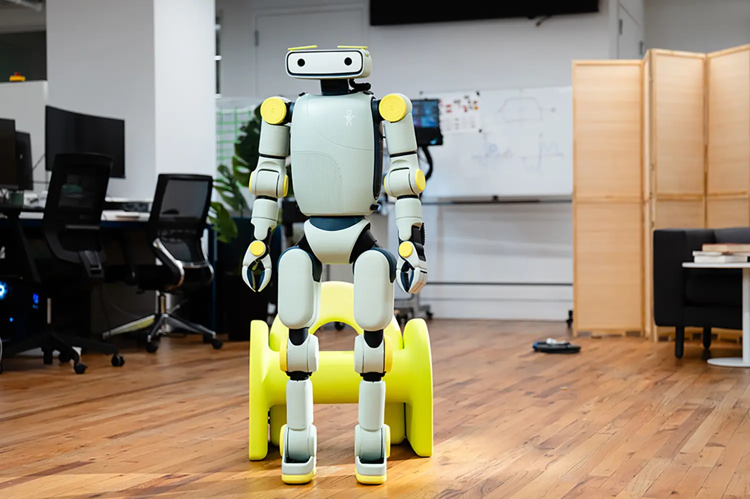 Fauna Robotics Sprout humanoid robot standing on yellow charging dock in office, front view, full body, mint-green and yellow palette, research lab background