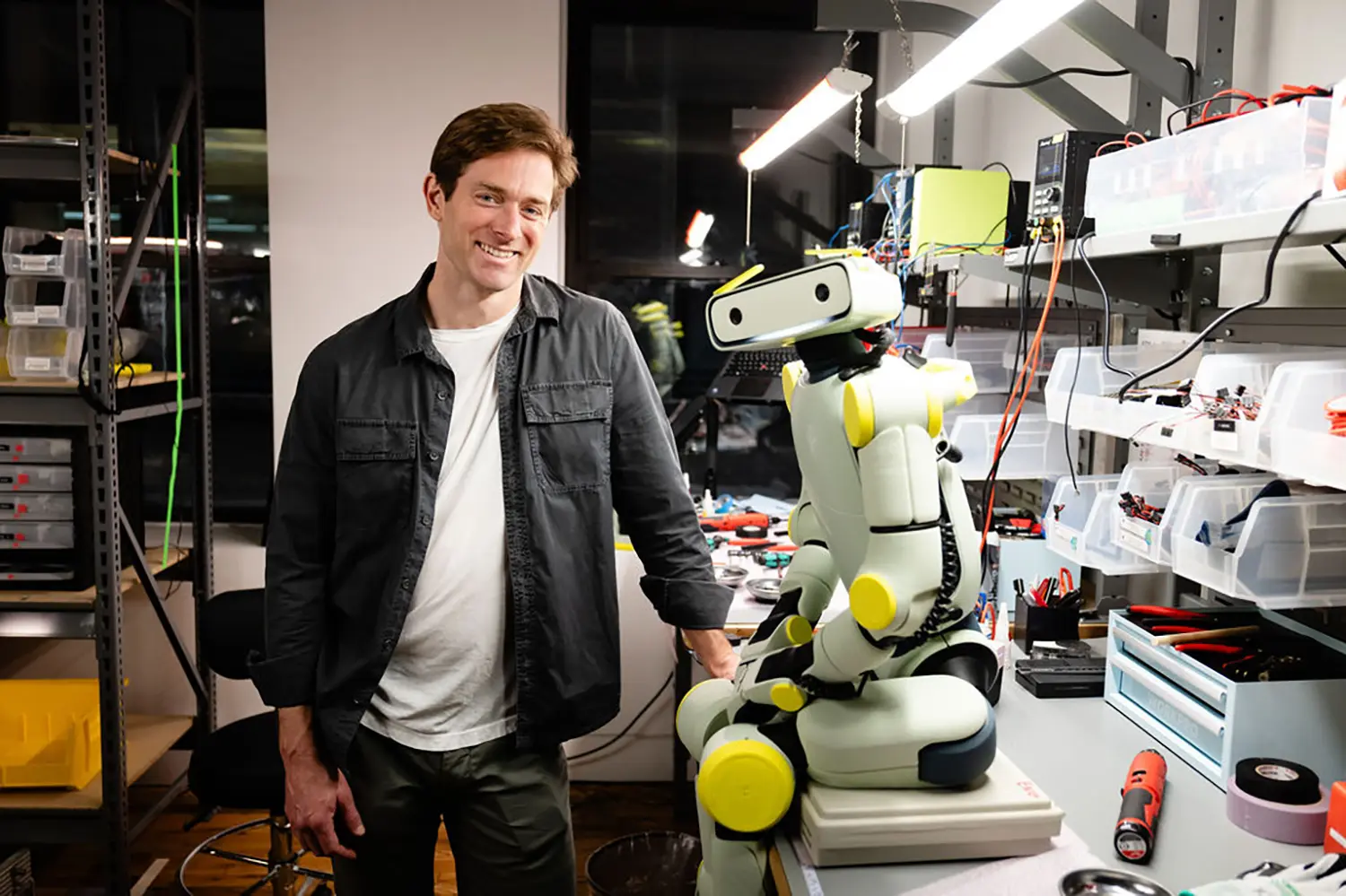 Fauna Robotics co-founder with Sprout humanoid robot on workbench, hardware lab setting, tools and components visible, robot in partial assembly