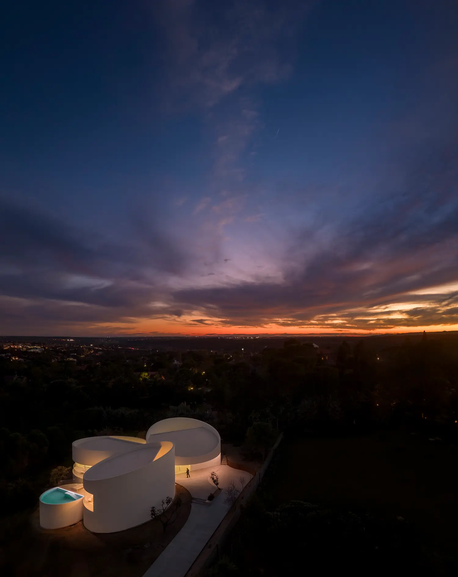 Twilight aerial view of House in Las Rozas, illuminated cylindrical volumes glowing against dark hillside and distant horizon