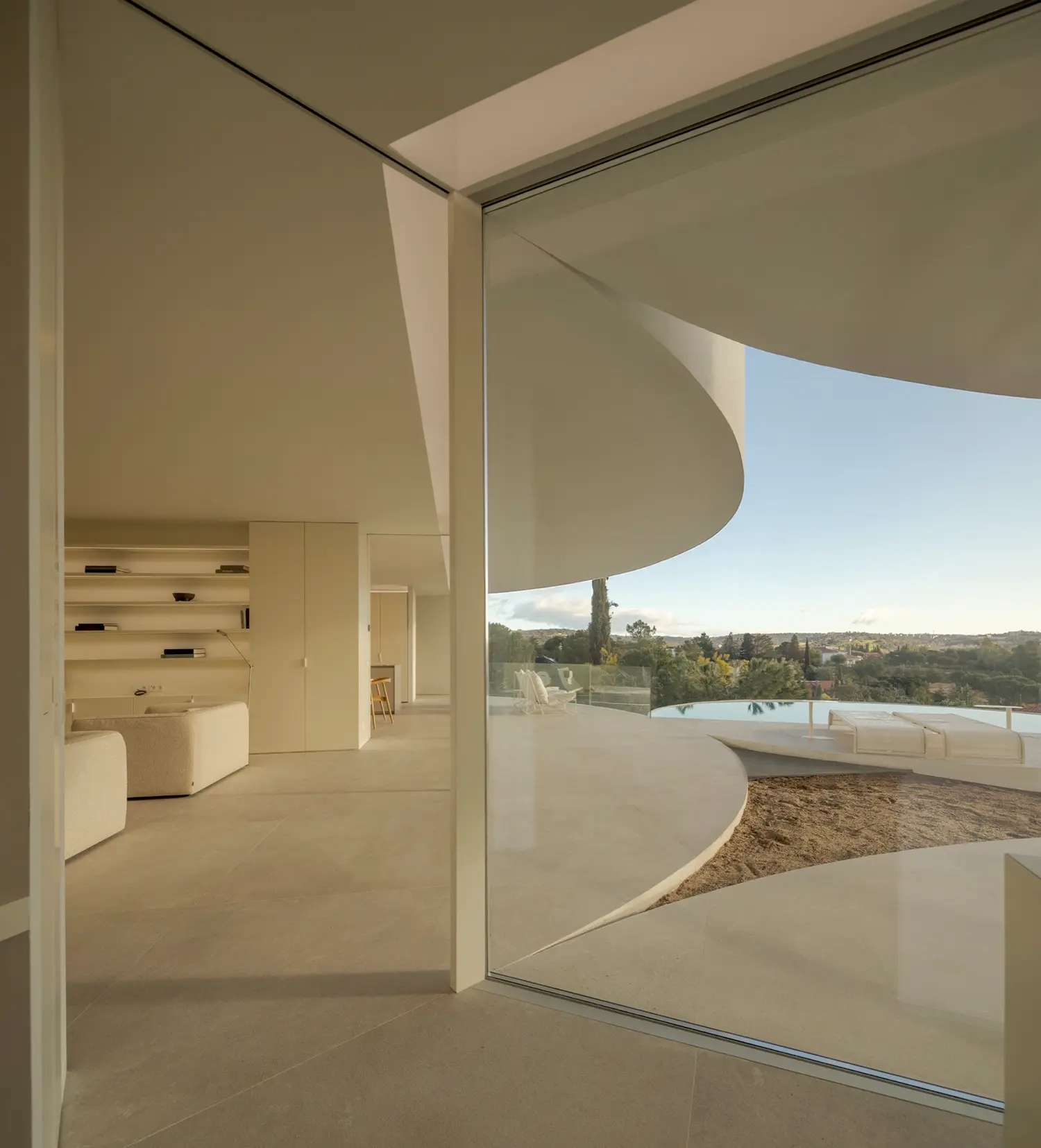 Living room interior of House in Las Rozas with curved upholstered seating, built-in shelving, and panoramic glazing toward landscape