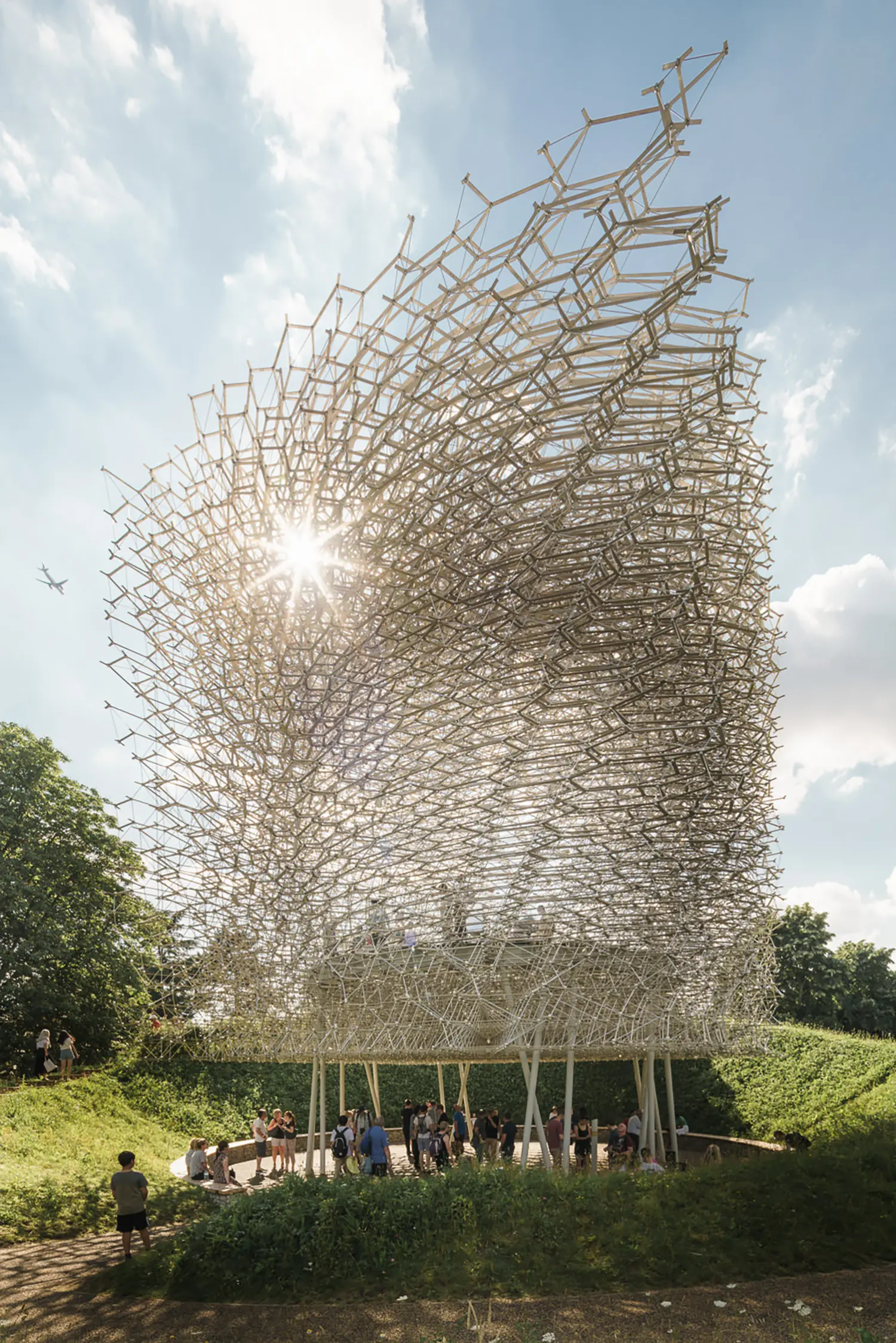 The Hive by Wolfgang Buttress at Kew Gardens, backlit aluminium lattice structure against blue sky with sunburst and visitors below