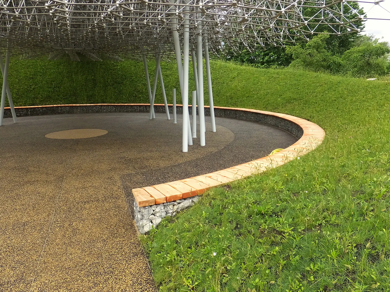 Gabion seating wall beneath The Hive by Wolfgang Buttress, wire cage filled with stone topped by timber capping, sloped meadow embankment behind