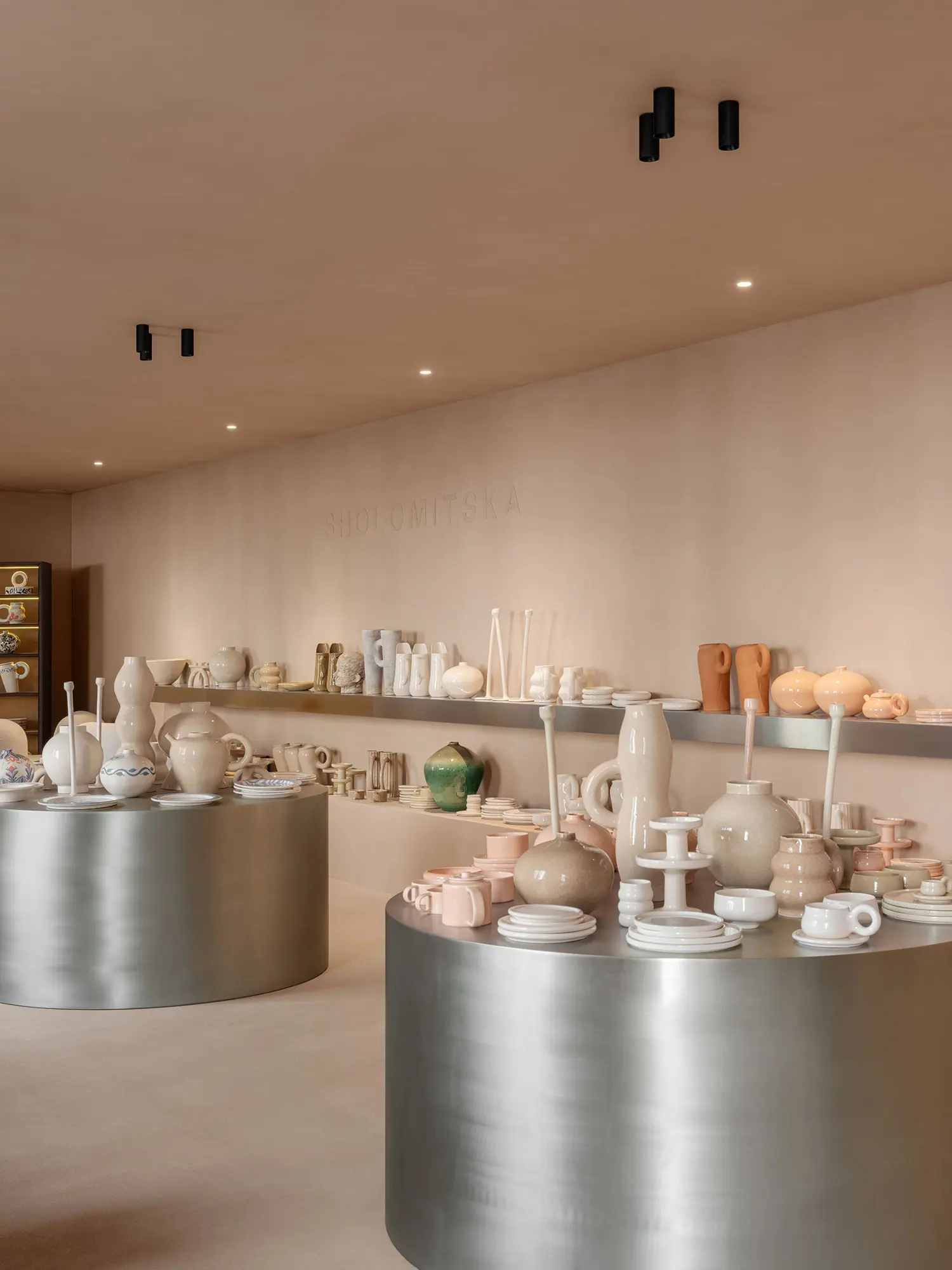 SHOLOMITSKA ceramics store Kyiv, retail area with brushed steel cylindrical plinths, handmade ceramic vessels and tableware, warm plaster walls, embossed brand lettering