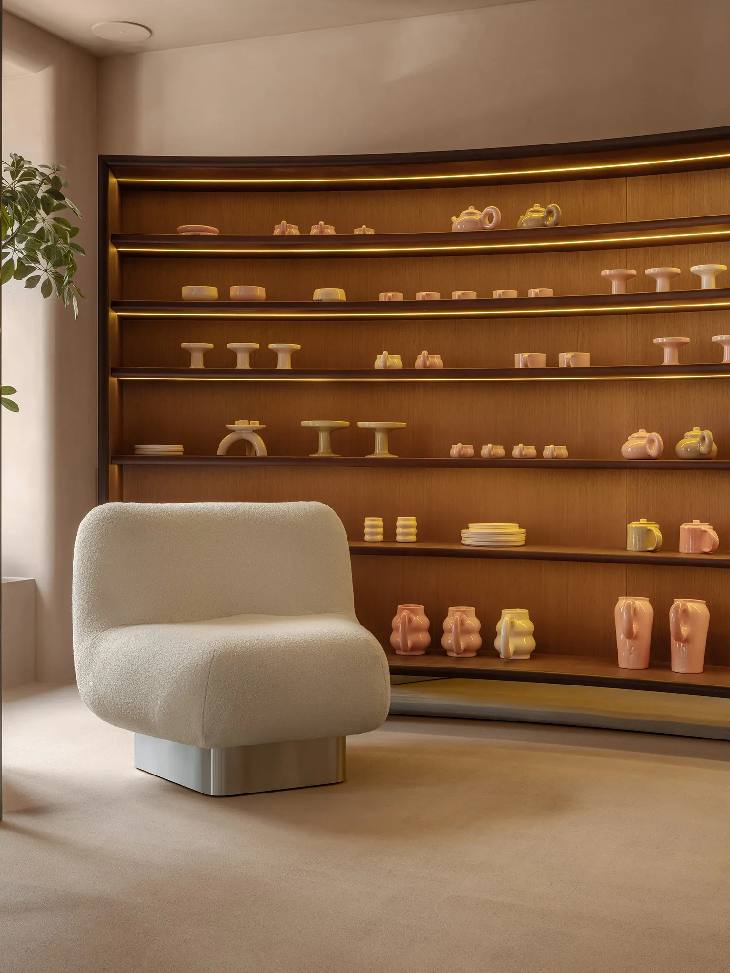SHOLOMITSKA ceramics store Kyiv, curved backlit wooden shelving unit with handmade pink and yellow ceramic pieces, bouclé lounge chair, warm ambient lighting
