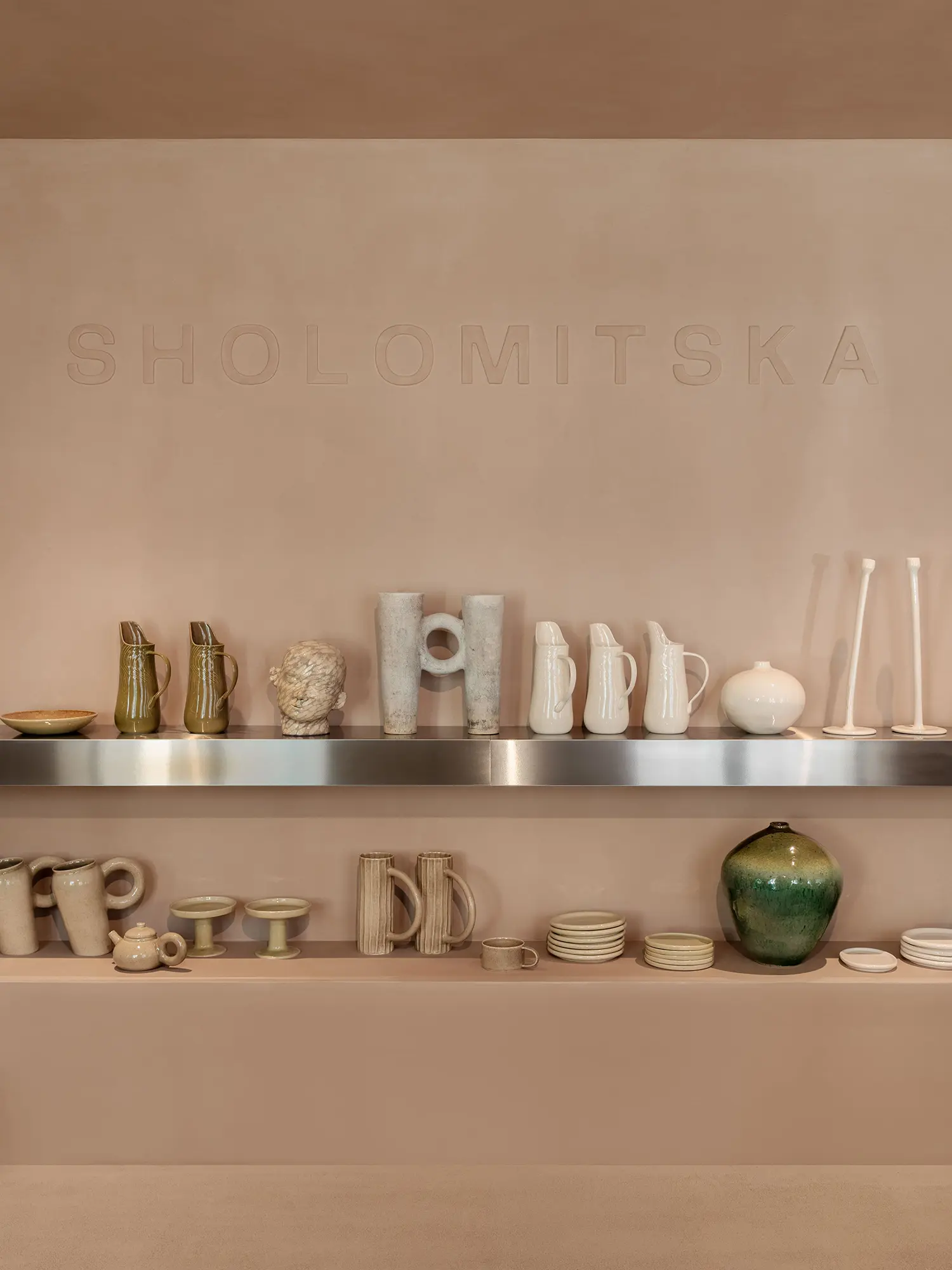 SHOLOMITSKA ceramics store Kyiv, wall shelves with handmade ceramic vessels jugs and tableware, embossed brand name in warm plaster wall, brushed steel shelf edge