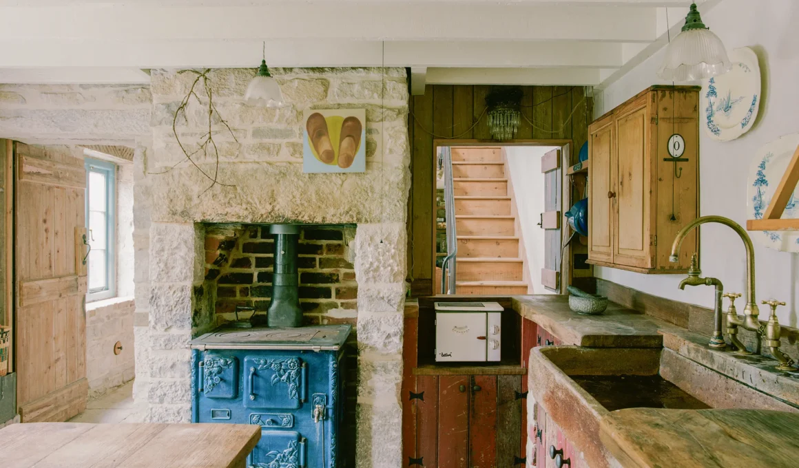 Kitchen at Purbeck Cottage by TYPE, restored blue cast-iron stove built into Purbeck stone chimney breast, reclaimed pine cabinetry, salvaged stone sink with brass taps